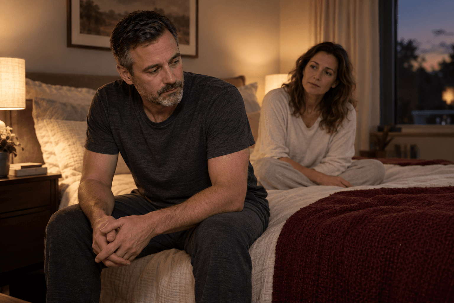 A man sits thoughtfully on the edge of the bed, with his partner behind him.